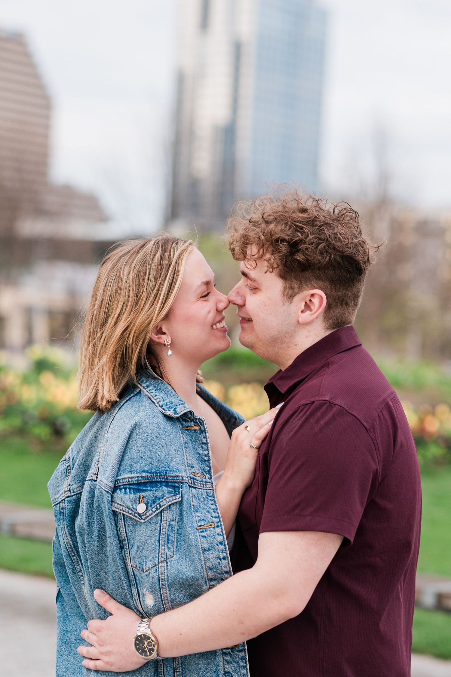 A romantic couple shares a tender nose-to-nose moment while embracing outdoors with a city skyline behind them.