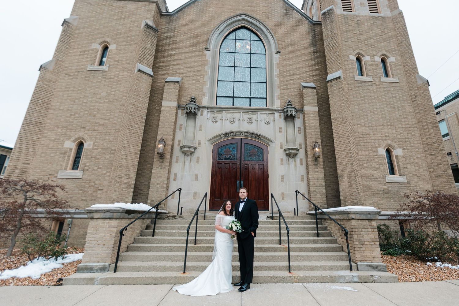 Bride and groom pose together on the steps of a grand stone church in winter wedding attire.