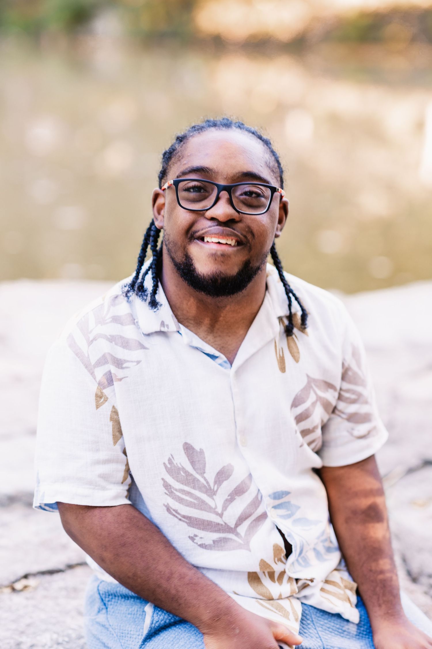 Smiling young man with dreadlocks and glasses sitting on rocks near water in a floral shirt.