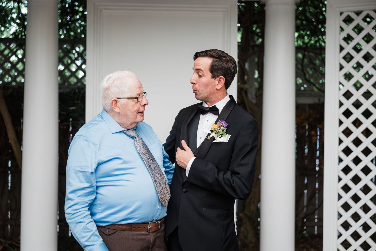 Groom in tuxedo with boutonniere sharing a conversation with an older man under a white lattice wedding gazebo.