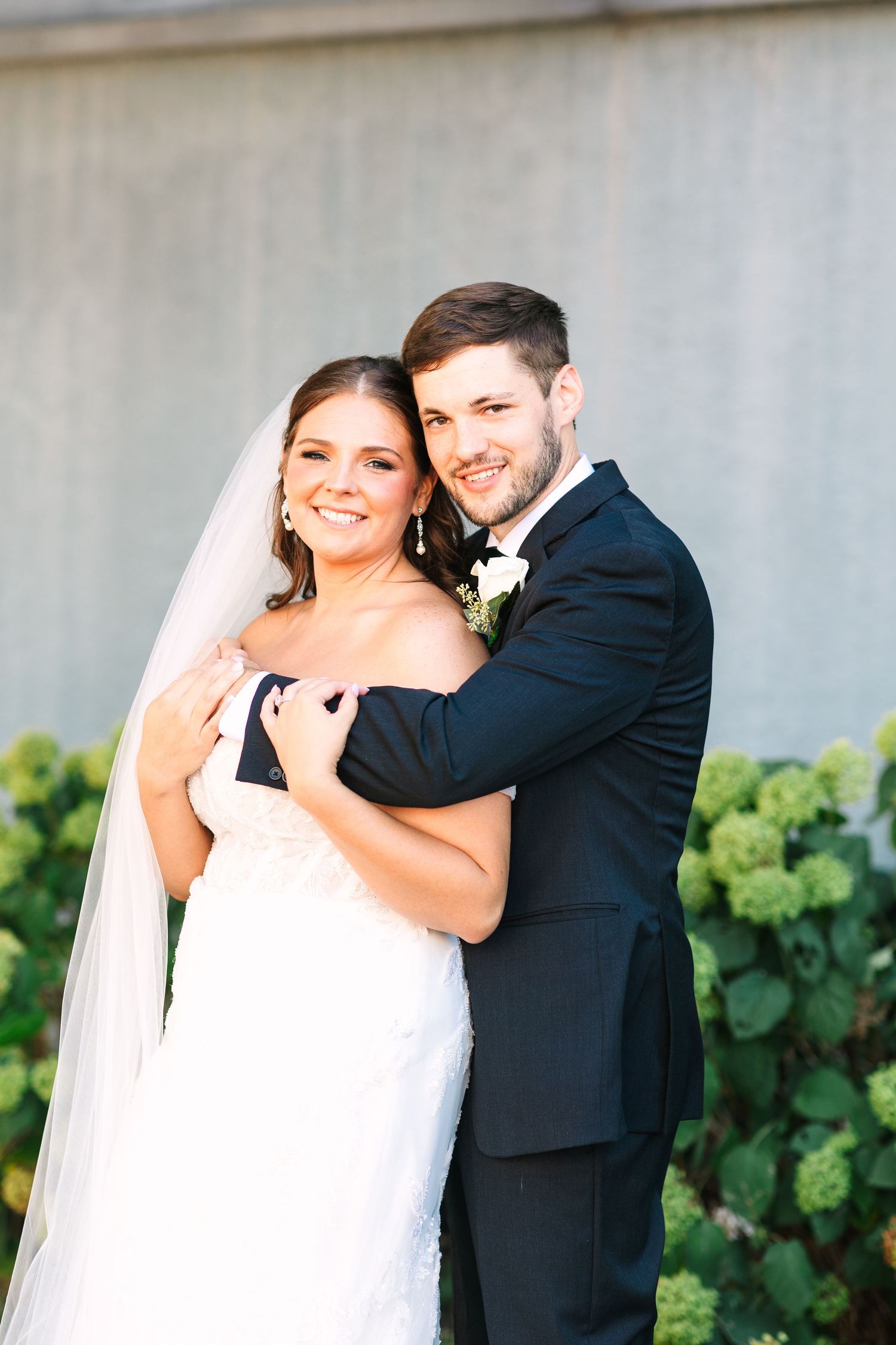Smiling bride in lace gown with veil embraced by groom in black suit before green hydrangea bushes.