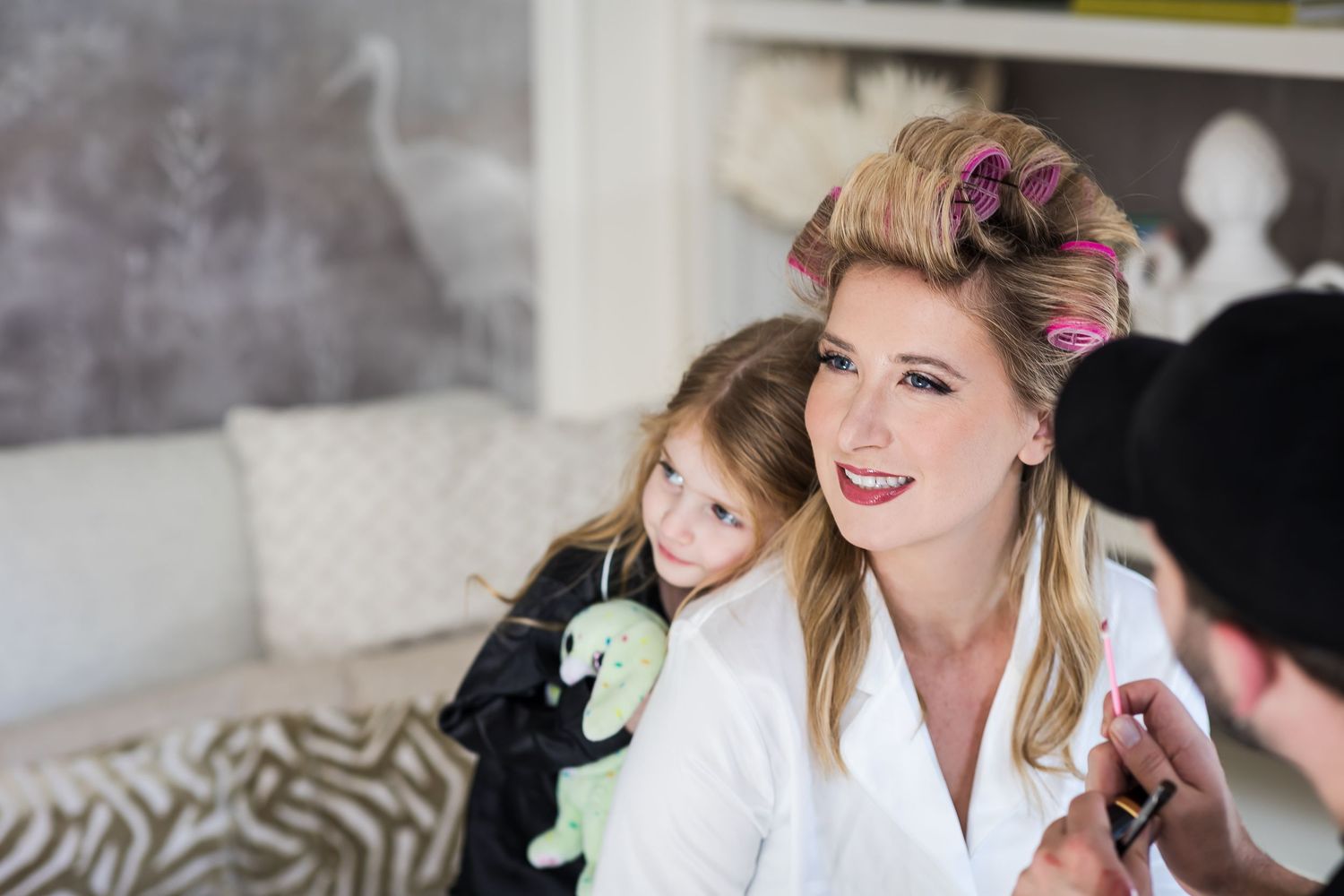 Woman in white robe with hair rollers smiles with young girl holding stuffed animal toy.