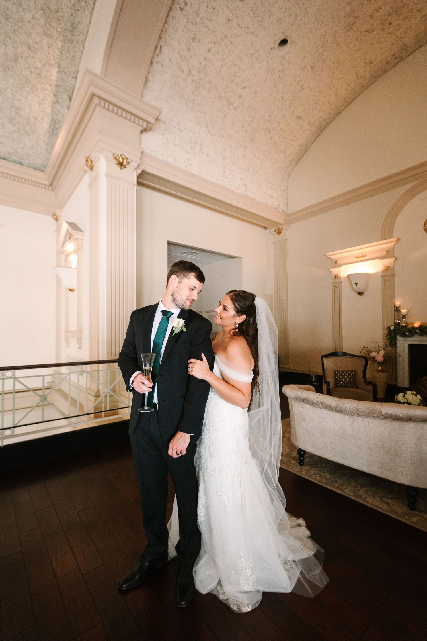 Bride in fitted lace gown and groom holding champagne sharing romantic moment in ornate classical interior.