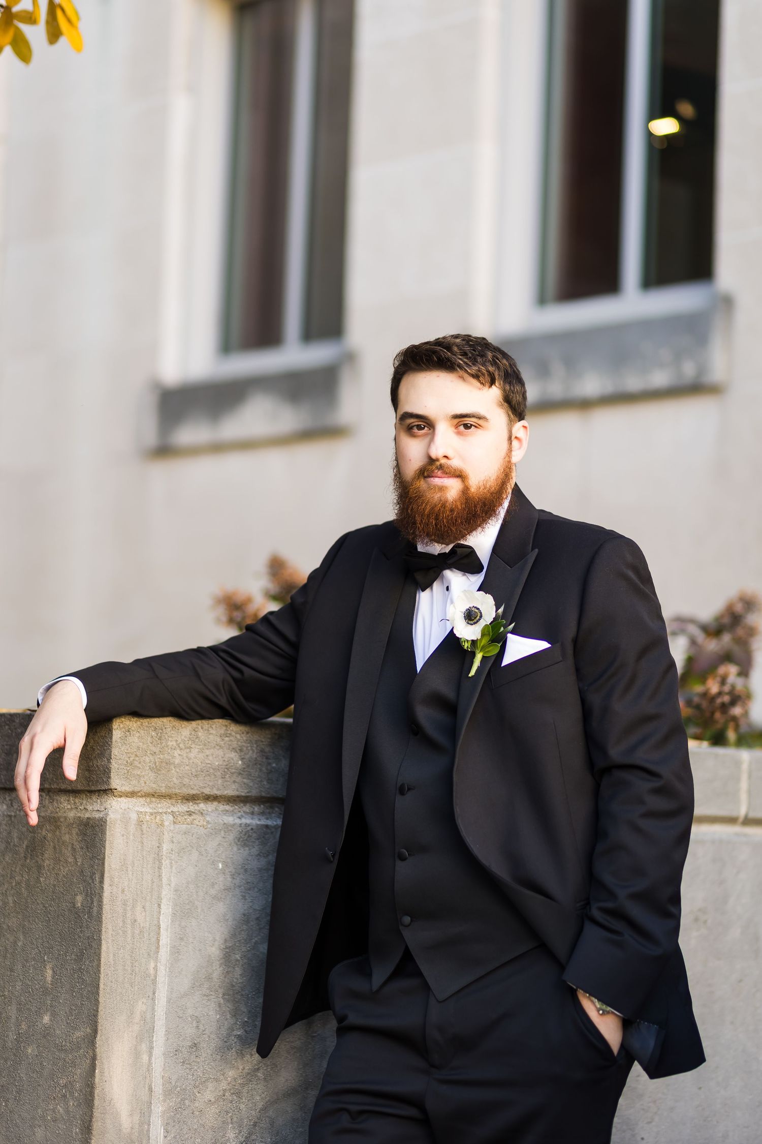 Bearded groom in black tuxedo with white boutonniere leans against stone wall outdoors the Cincinnati Art Museum.