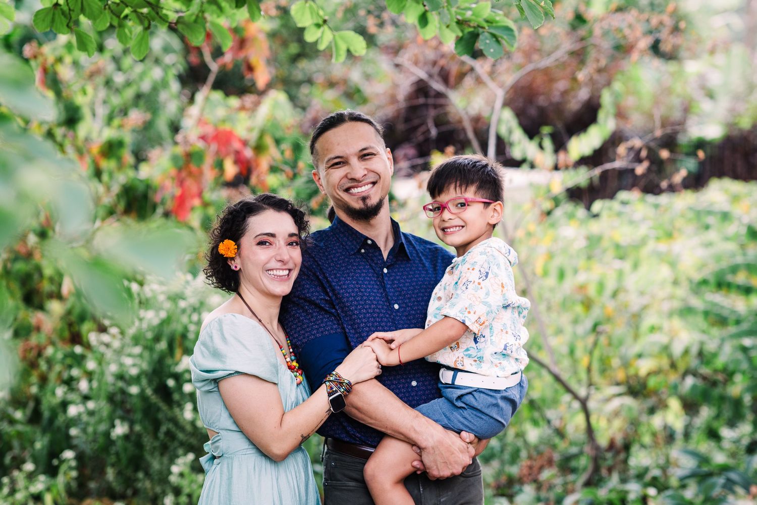 Smiling family of three with young son posing in tropical garden setting.