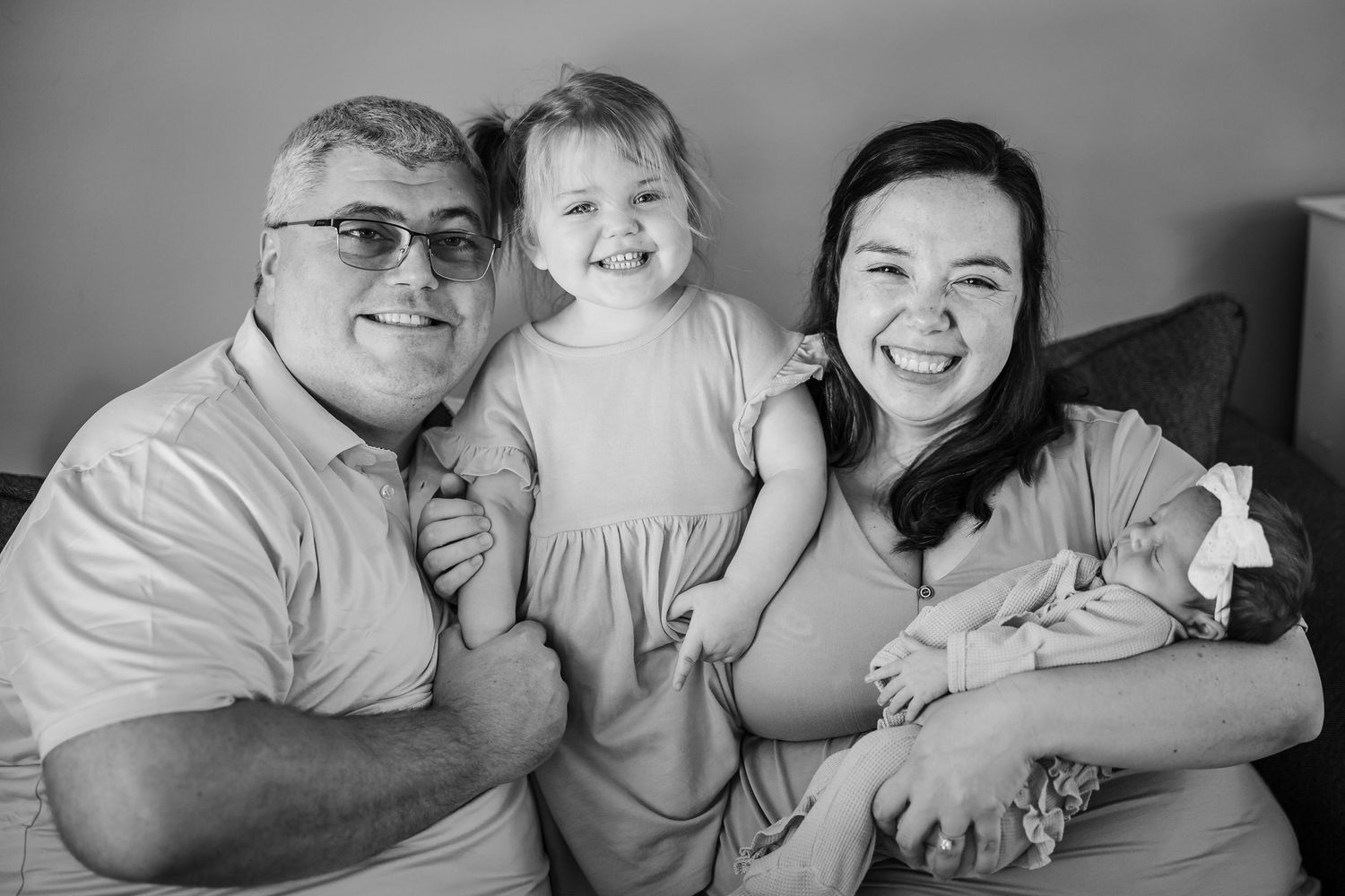 A black and white family portrait of parents laughing with their toddler daughter and newborn baby.