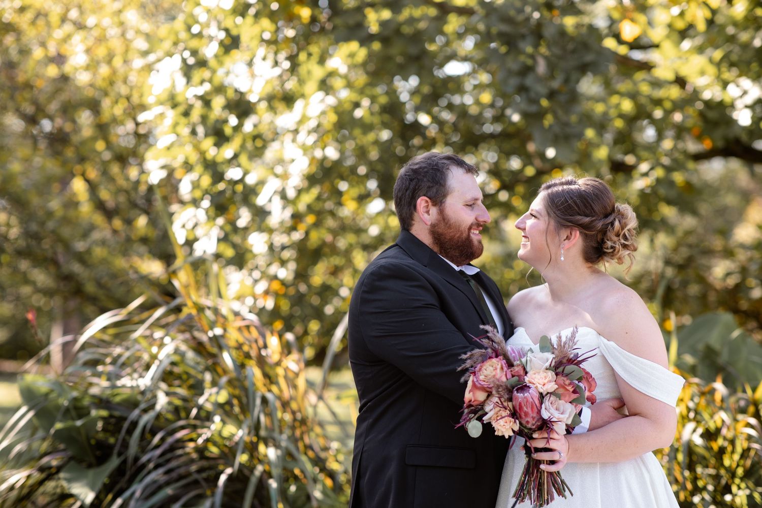 Bride and groom share romantic moment outdoors surrounded by lush golden foliage holding floral bouquet.