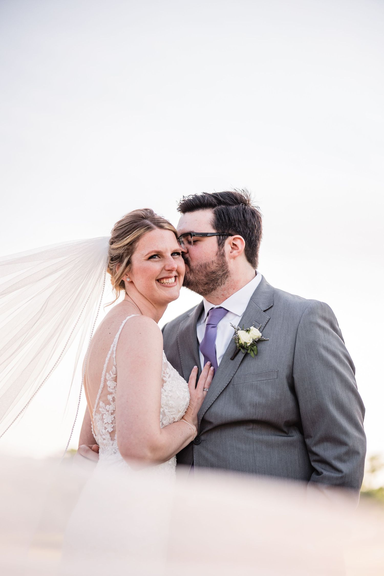 Groom kisses bride on cheek as her veil blows in the wind against a bright sky.