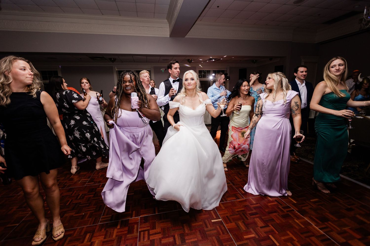 Bride in white ball gown dancing energetically with bridesmaids in lavender dresses at a wedding reception at the Manor House.
