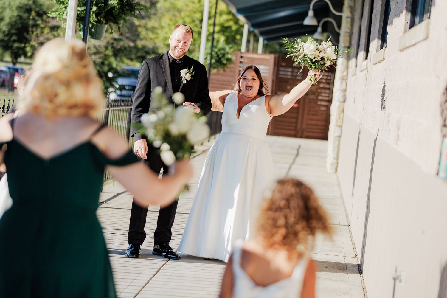 Bride in white gown joyfully greets guests on a sunny outdoor walkway with groom nearby at Lake Lyndsay.
