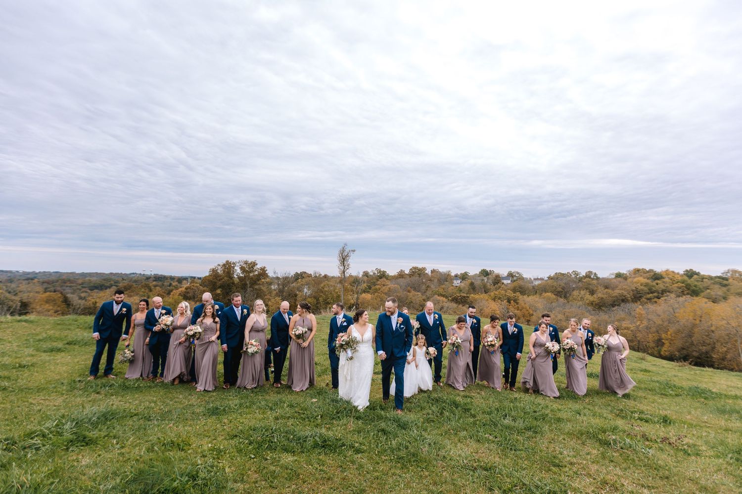 Large wedding party in navy and taupe standing in a scenic green countryside field.