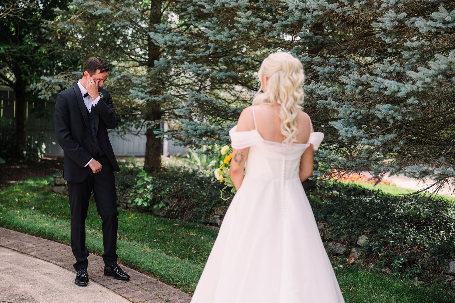Emotional groom in black suit reacts seeing blonde bride in white off-shoulder gown holding yellow bouquet outdoors.