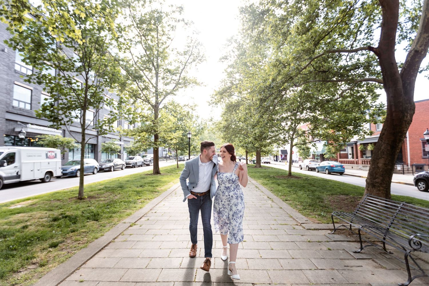 Couple walking and smiling together along a tree-lined urban sidewalk on a bright sunny day.