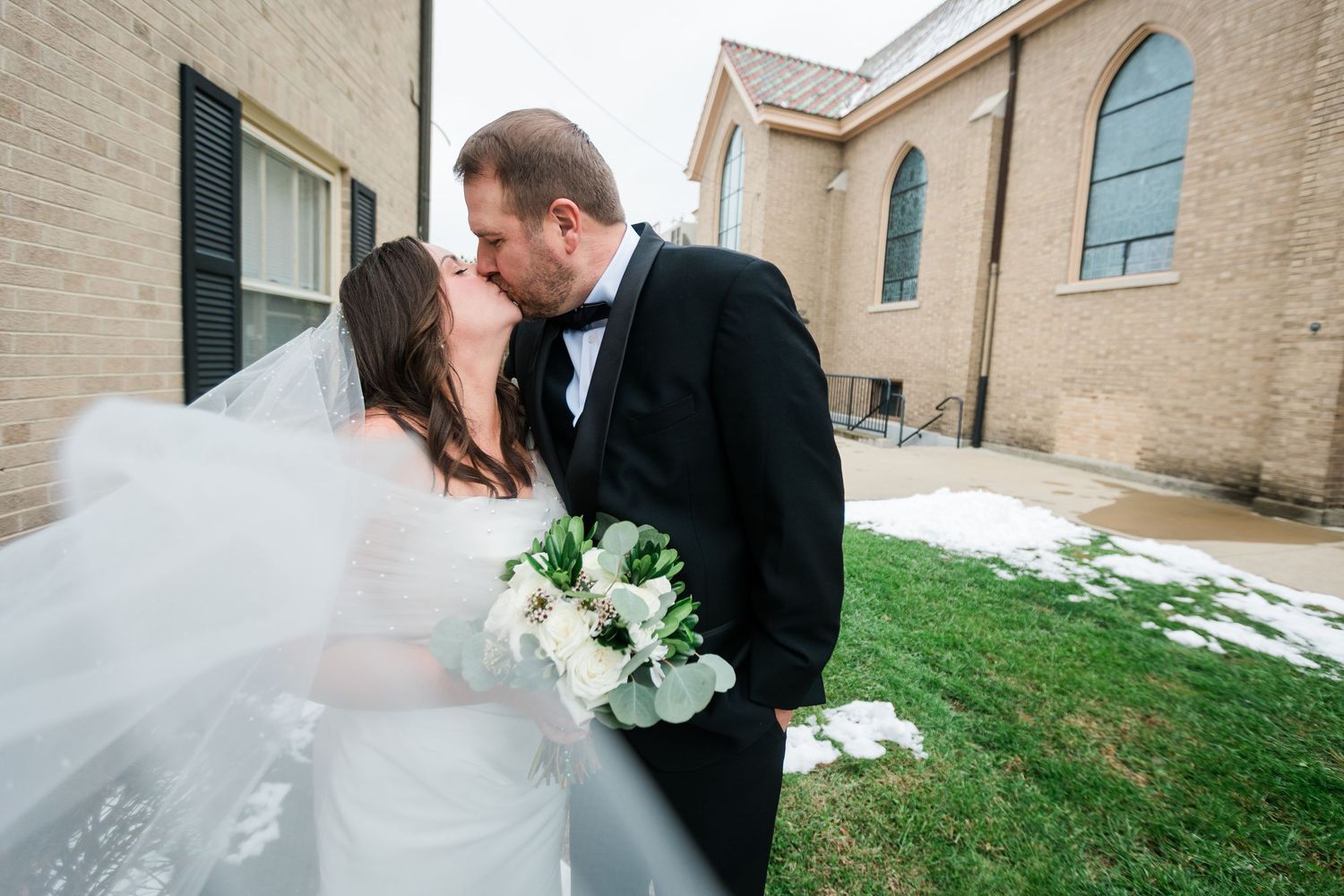 Bride and groom share a romantic kiss outside a brick church, with the bride's veil flowing in the wind.
