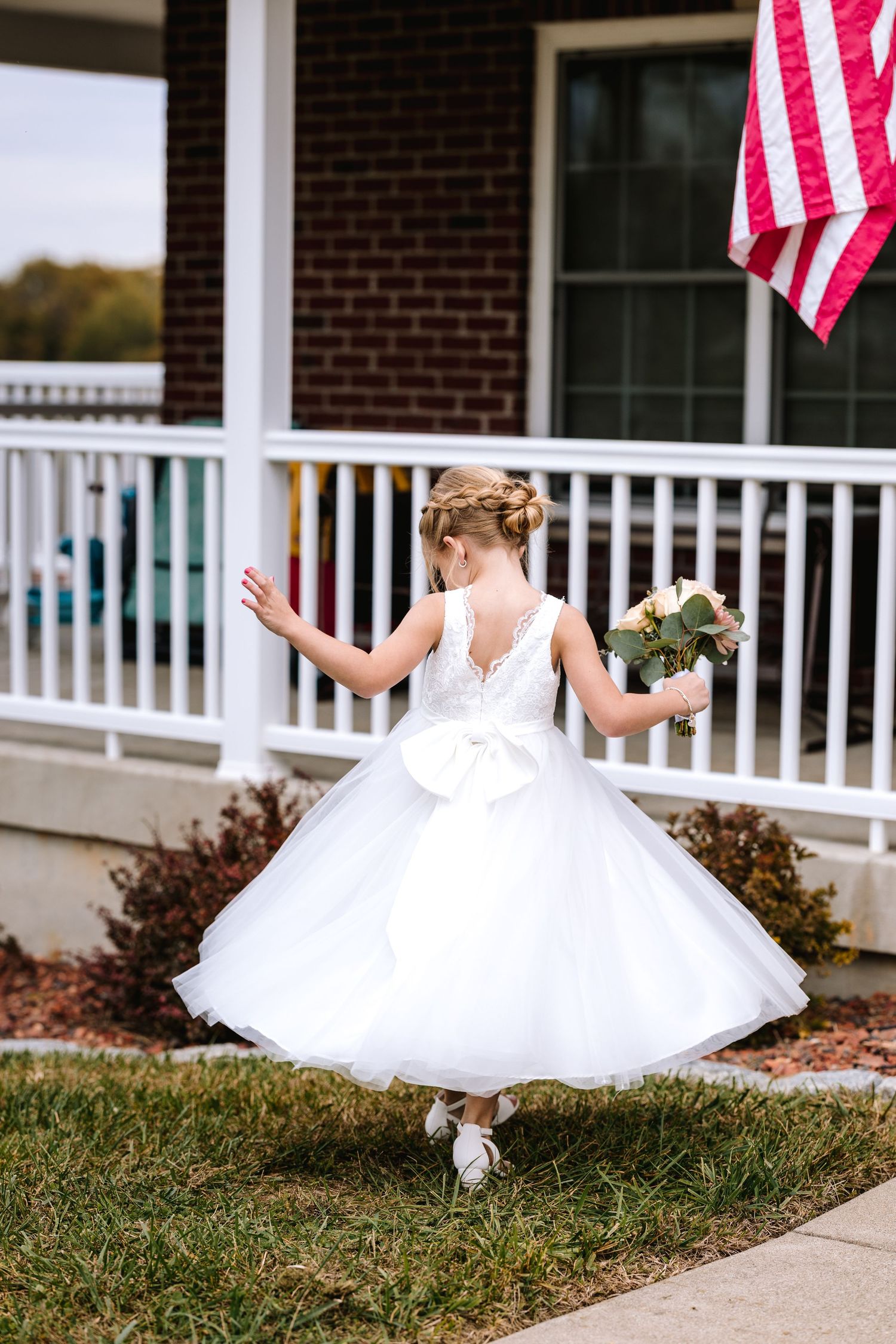 Flower girl in white tulle dress twirling with bouquet near a brick porch.
