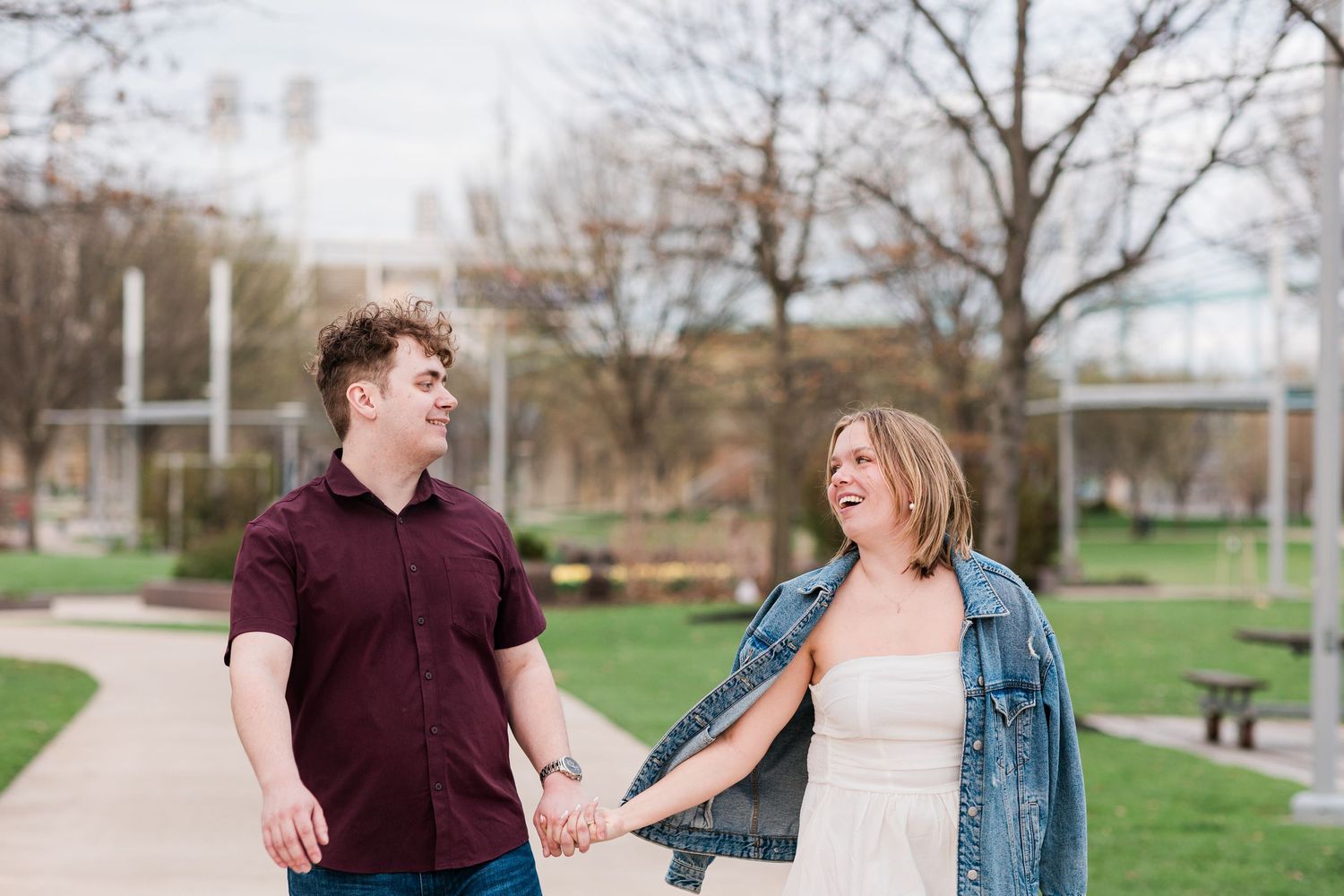 A happy couple holds hands and laughs while walking through a park with city buildings in the background.
