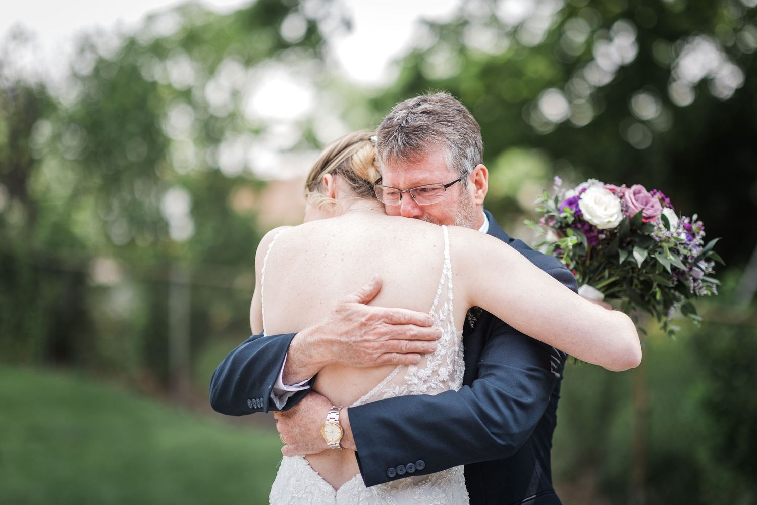 Bride and father share an emotional hug outdoors, with bridal bouquet visible during wedding day first look.