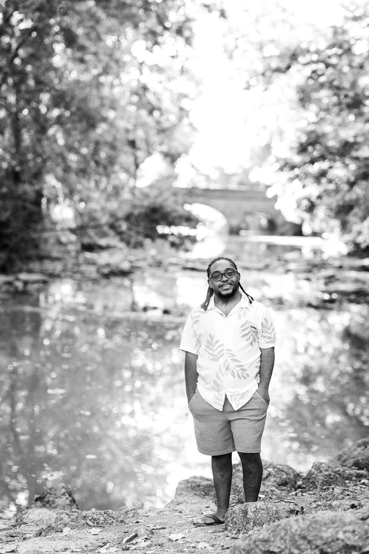 Smiling man with dreadlocks and glasses standing by a reflective pond outdoors, black and white photo.