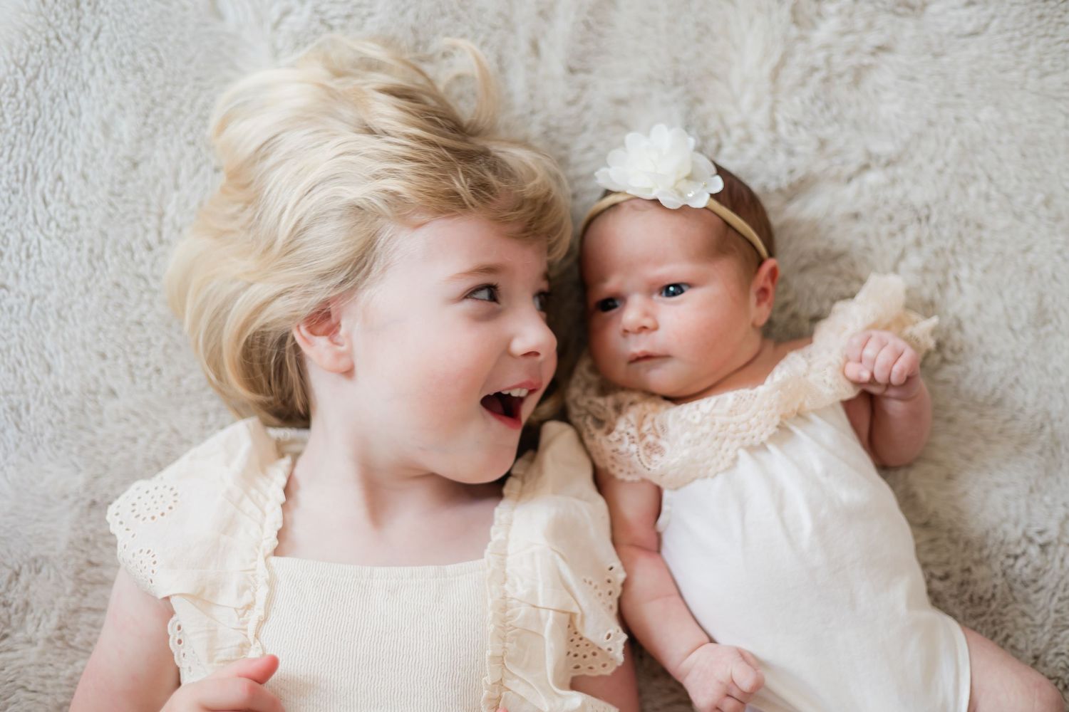 Young girl with big smile lying next to newborn baby sister in matching cream outfits.