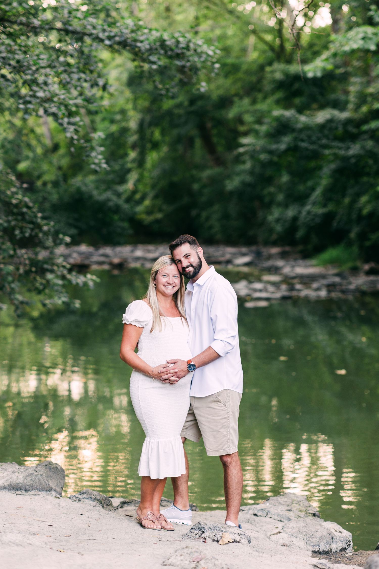 Pregnant couple embracing by a serene green pond surrounded by lush trees in summer at Sharon Woods.