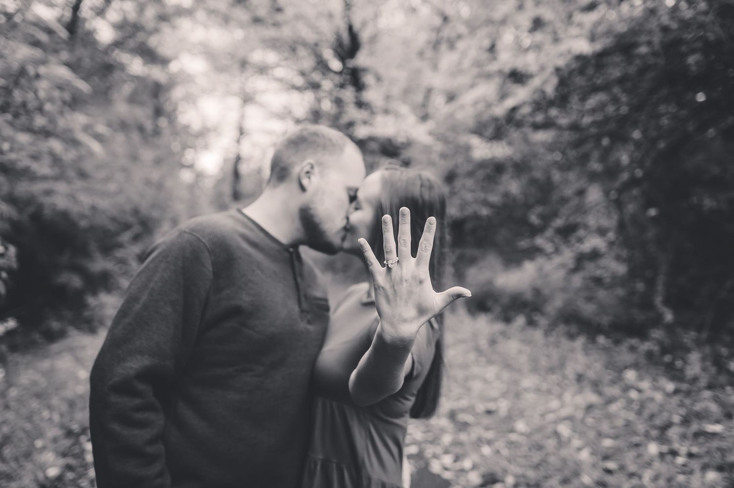 Black and white photo of couple kissing in forest while showing engagement ring.