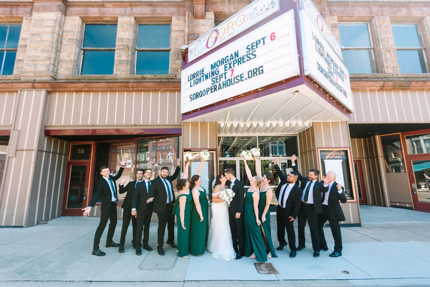 Wedding party in emerald green and black attire celebrating outside vintage theater marquee sign at dusk.