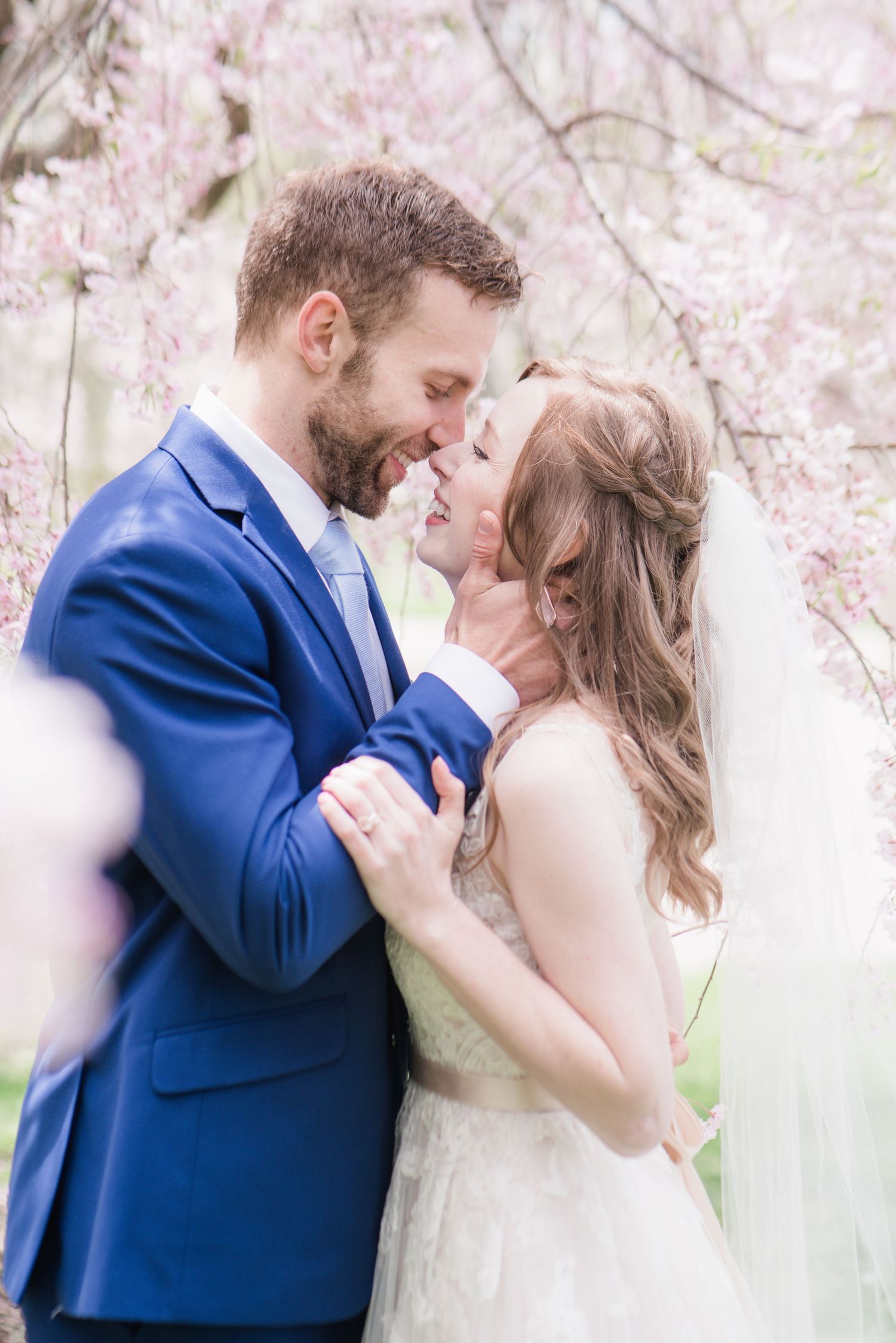 Couple embracing and smiling under blooming pink cherry blossoms in romantic wedding portrait.