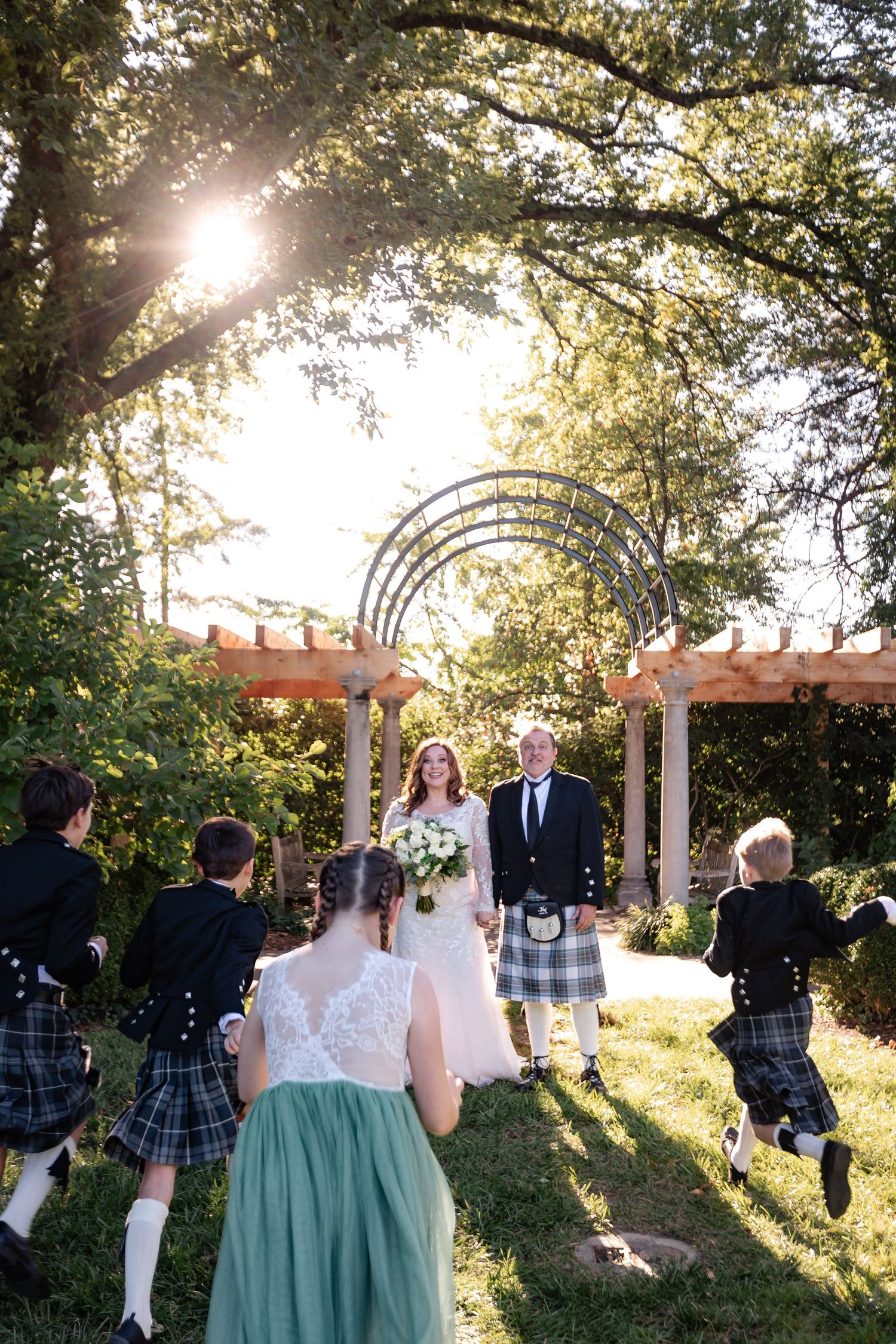 Bride and groom in Scottish kilts greeted by children running toward them at sunset.