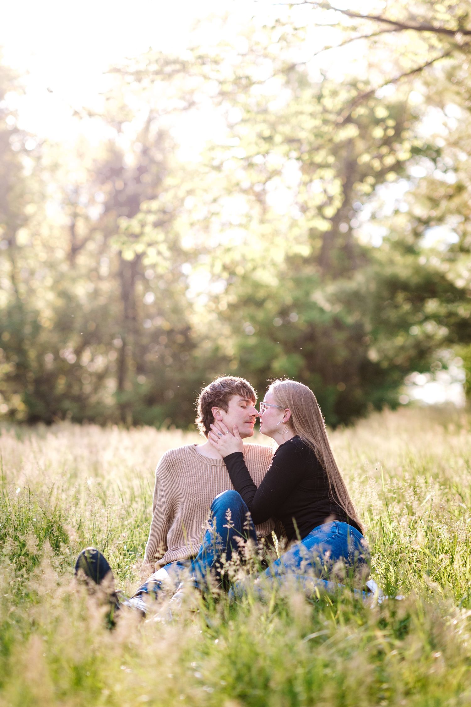 A young couple shares a romantic kiss while sitting together in a sunlit meadow with tall grass.
