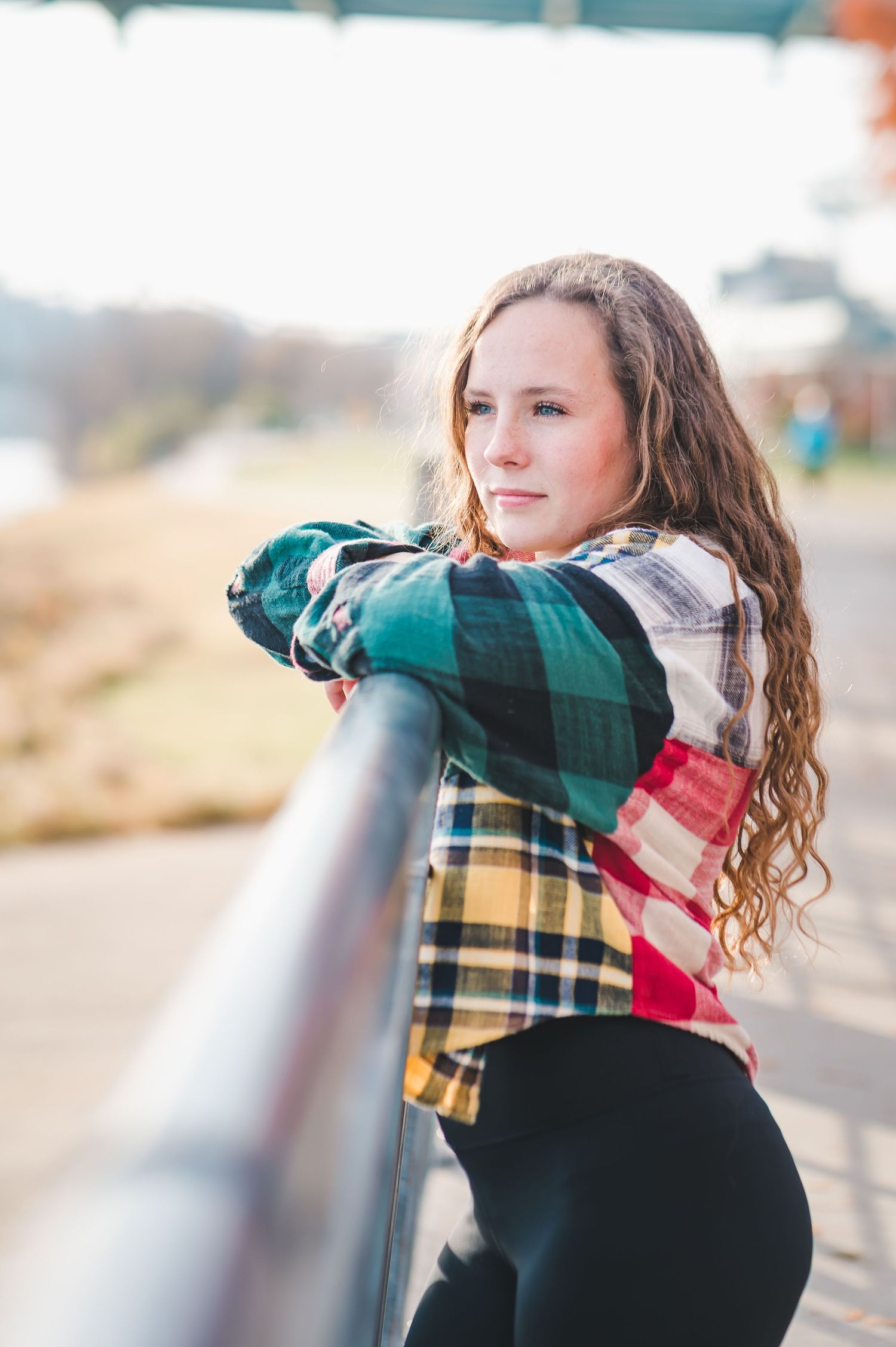 Young woman in multicolor plaid shirt leaning on metal railing looking over shoulder.