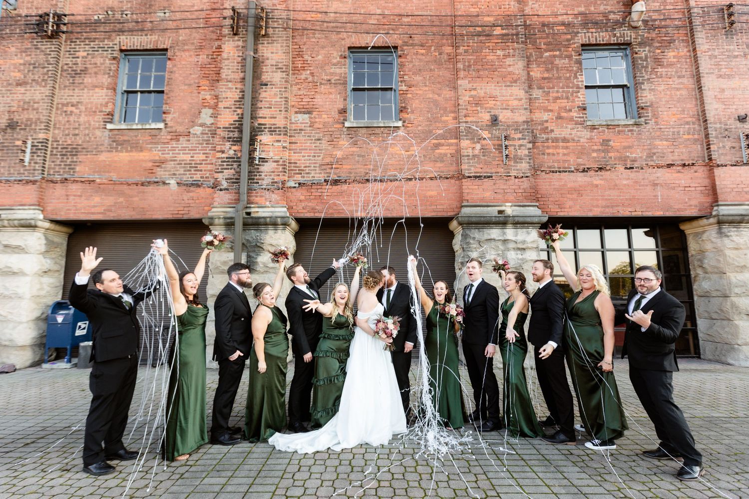 Wedding party in green dresses and black suits celebrating outside a brick building with champagne streamers.