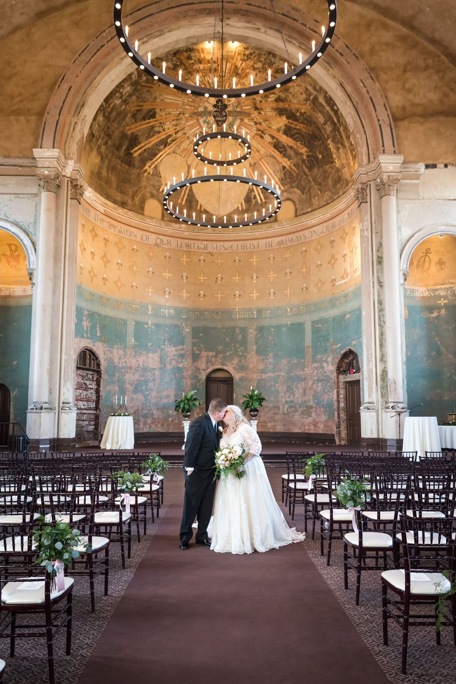 Bride and groom sharing a kiss in an ornate wedding venue with arched ceilings and elegant chandelier.