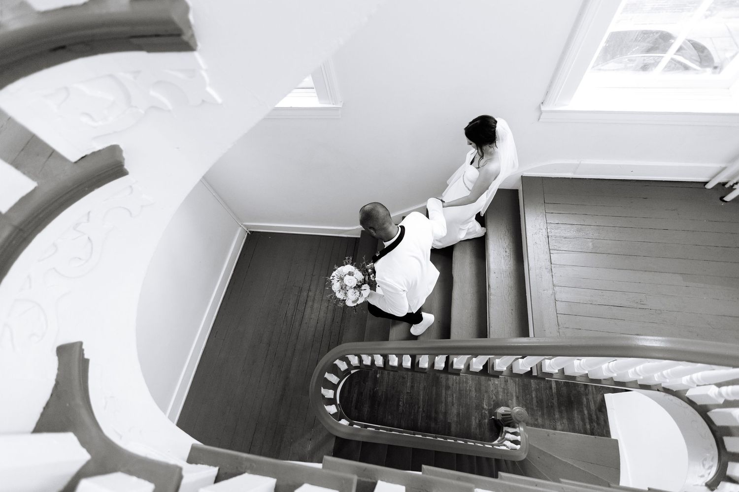 Aerial black and white view of bride and groom descending an elegant spiral staircase together.