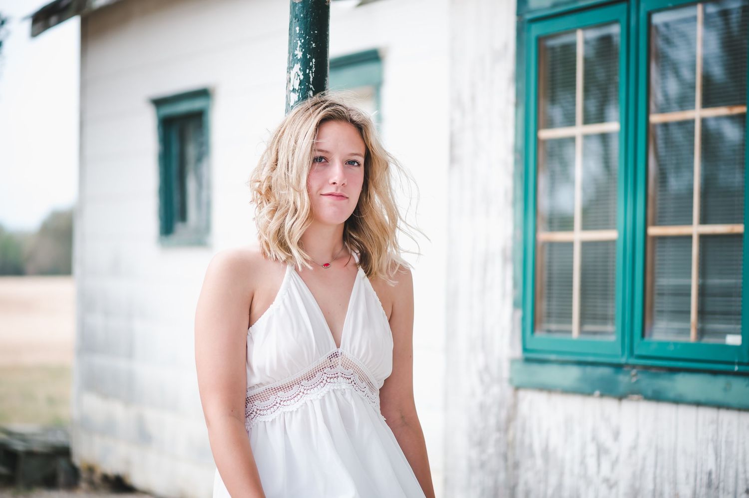 Young blonde woman in a white halter dress stands near a vintage building with teal-trimmed windows.