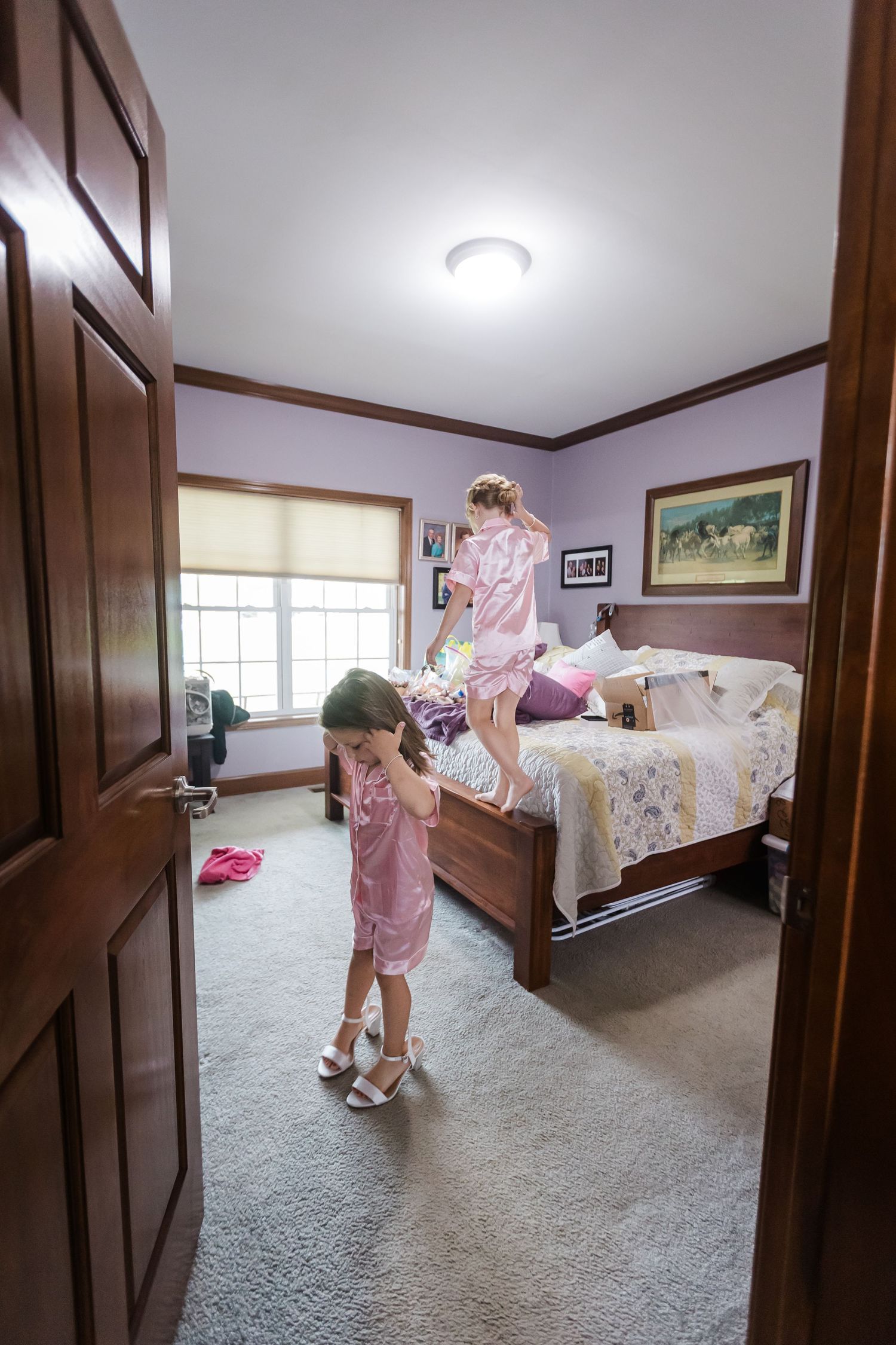 Two young flower girls in pink robes play in a bedroom, one jumping on the bed before the wedding.