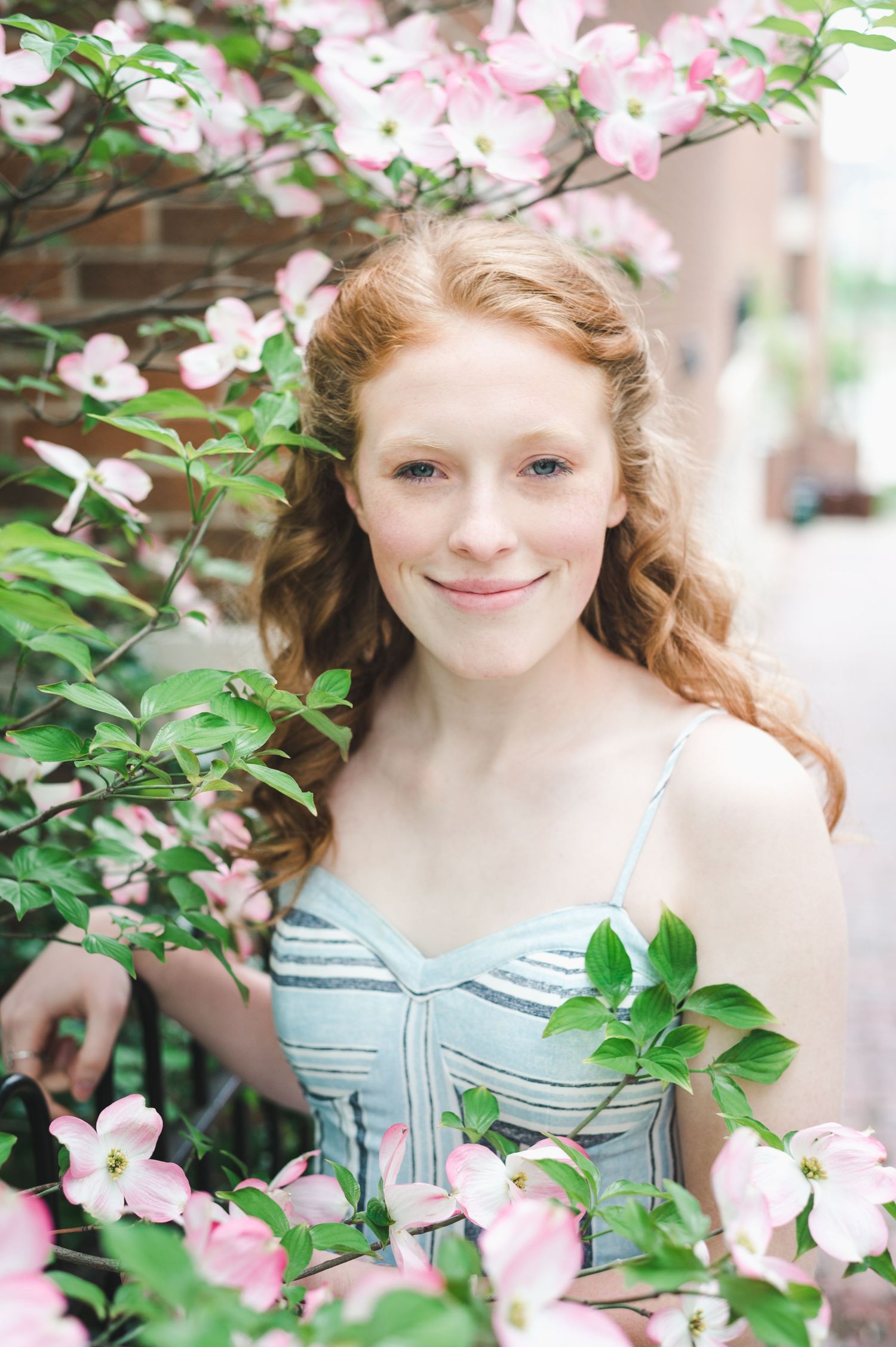 Smiling young woman with red hair stands among blooming pink dogwood flowers in a blue dress.