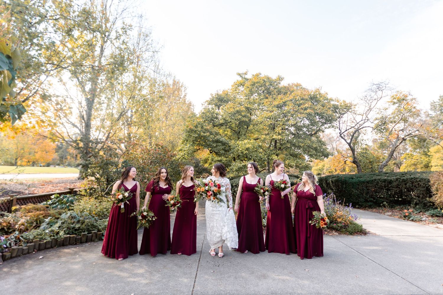 Bride in white lace gown walks with bridesmaids in burgundy dresses through autumn garden setting.