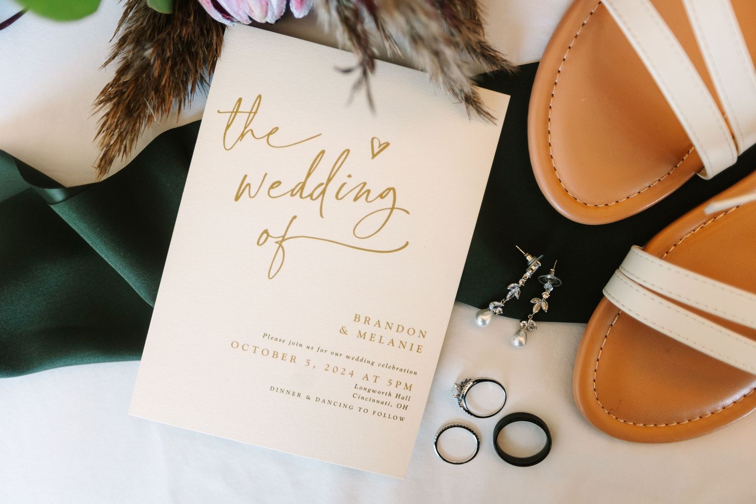 Wedding program for Brandon and Melanie with gold script lettering, rings, earrings, and sandals on a green cloth.