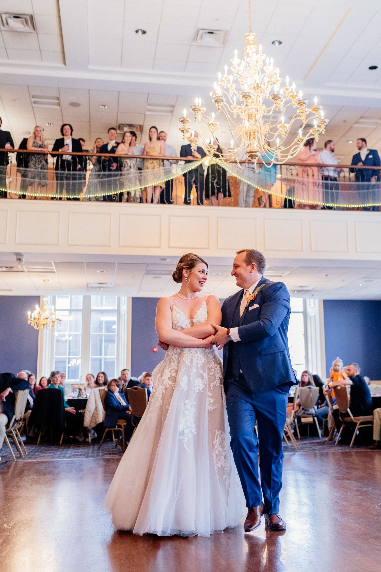 Bride and groom share their first dance under a grand chandelier as guests watch from a balcony.