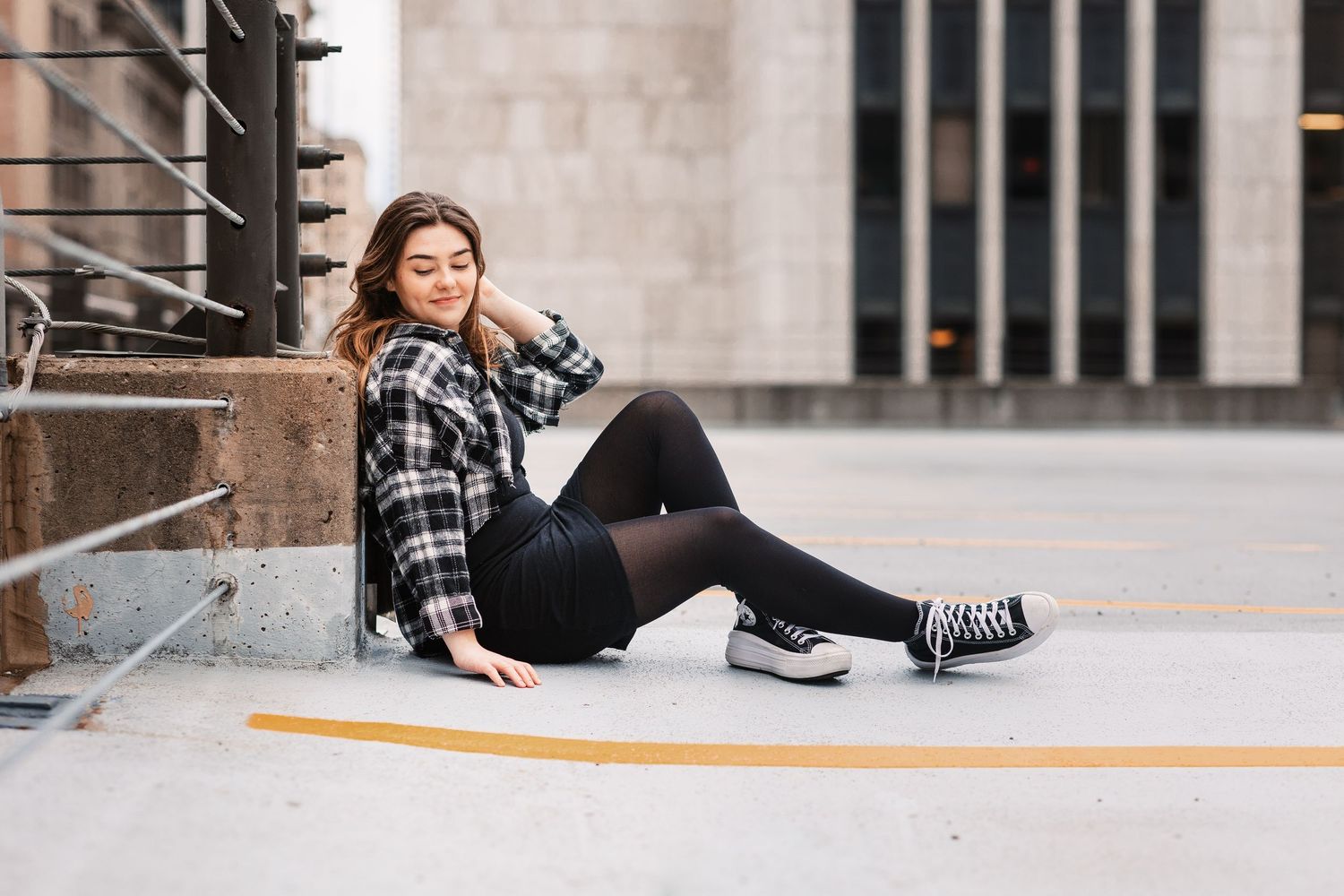 Young woman in plaid shirt and black leggings sitting on urban pavement against building wall.