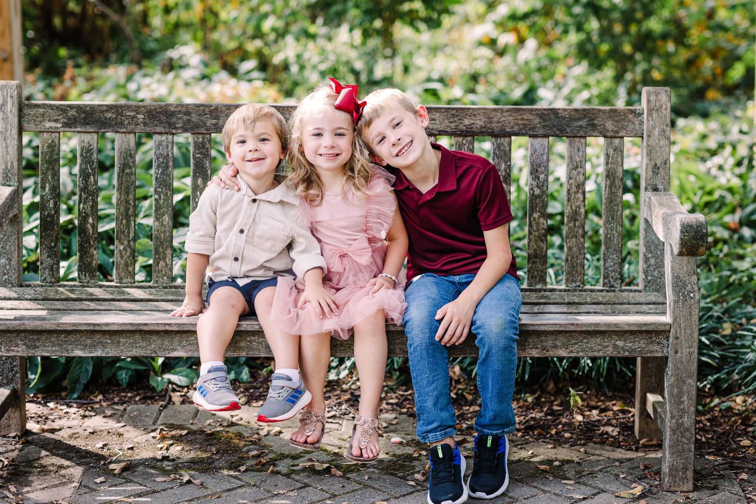 Three young siblings sitting on a wooden bench outdoors, smiling together in a garden setting.