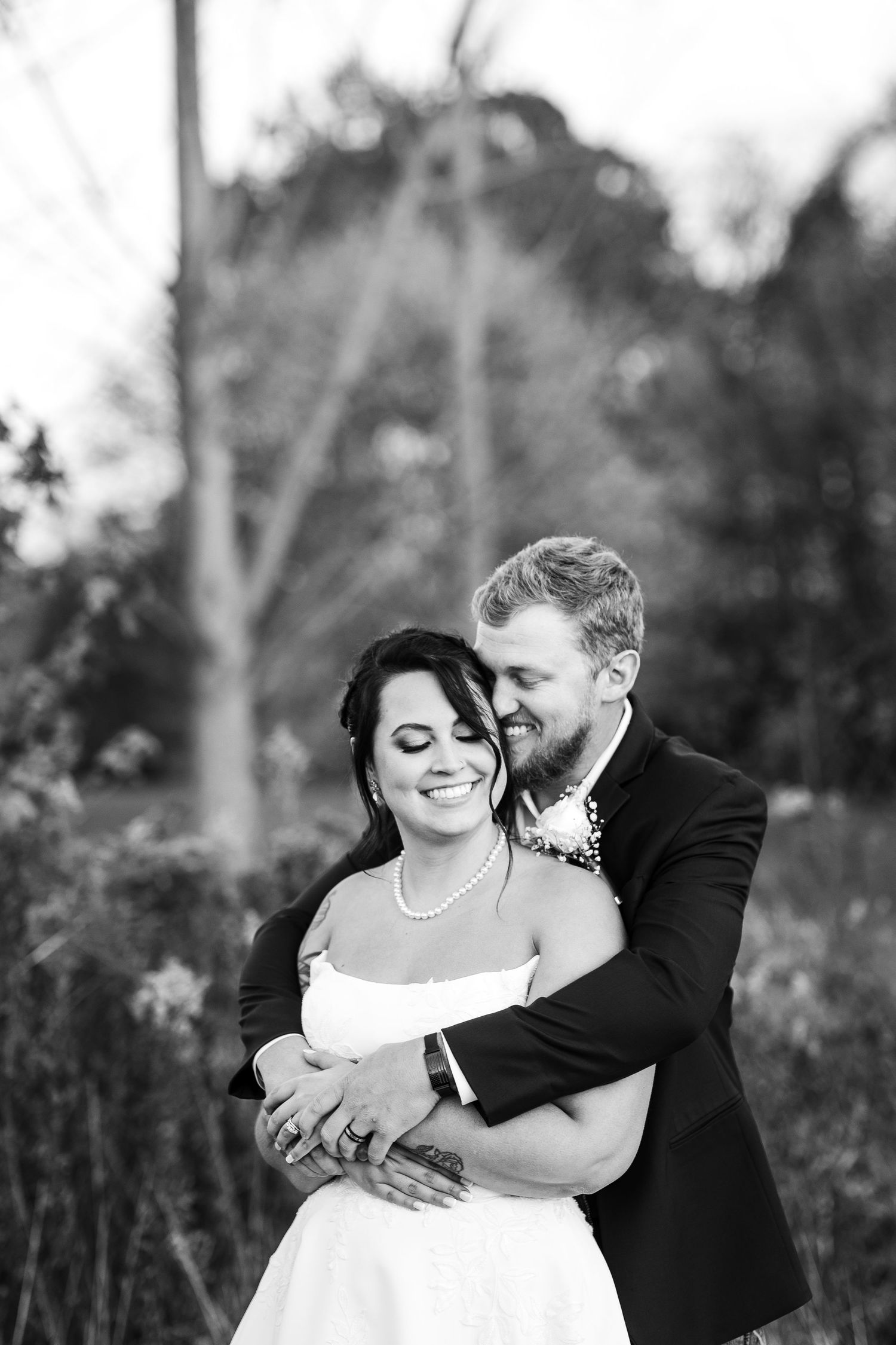 Black and white photo of groom embracing bride from behind in a romantic outdoor wedding portrait.