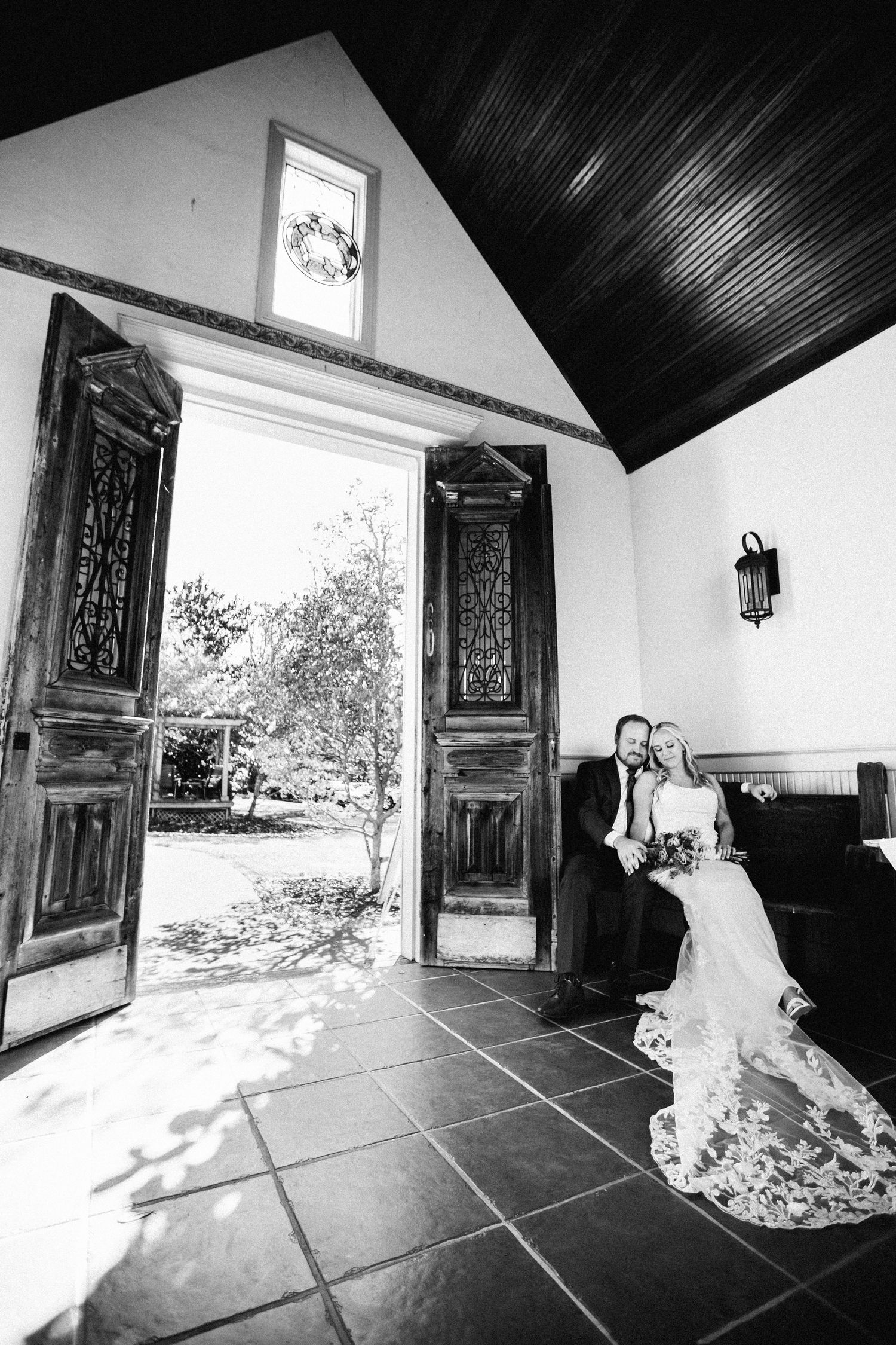 Black and white photo of bride and groom sitting near ornate open chapel doors at the Inn at Oneonta.