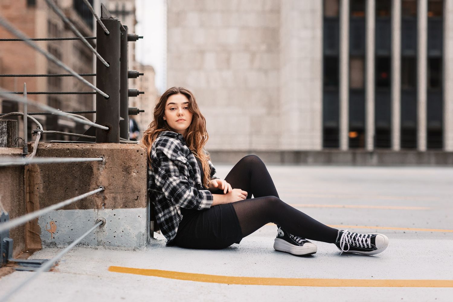 Teenage girl in plaid shirt and black leggings sitting on urban steps with city background.