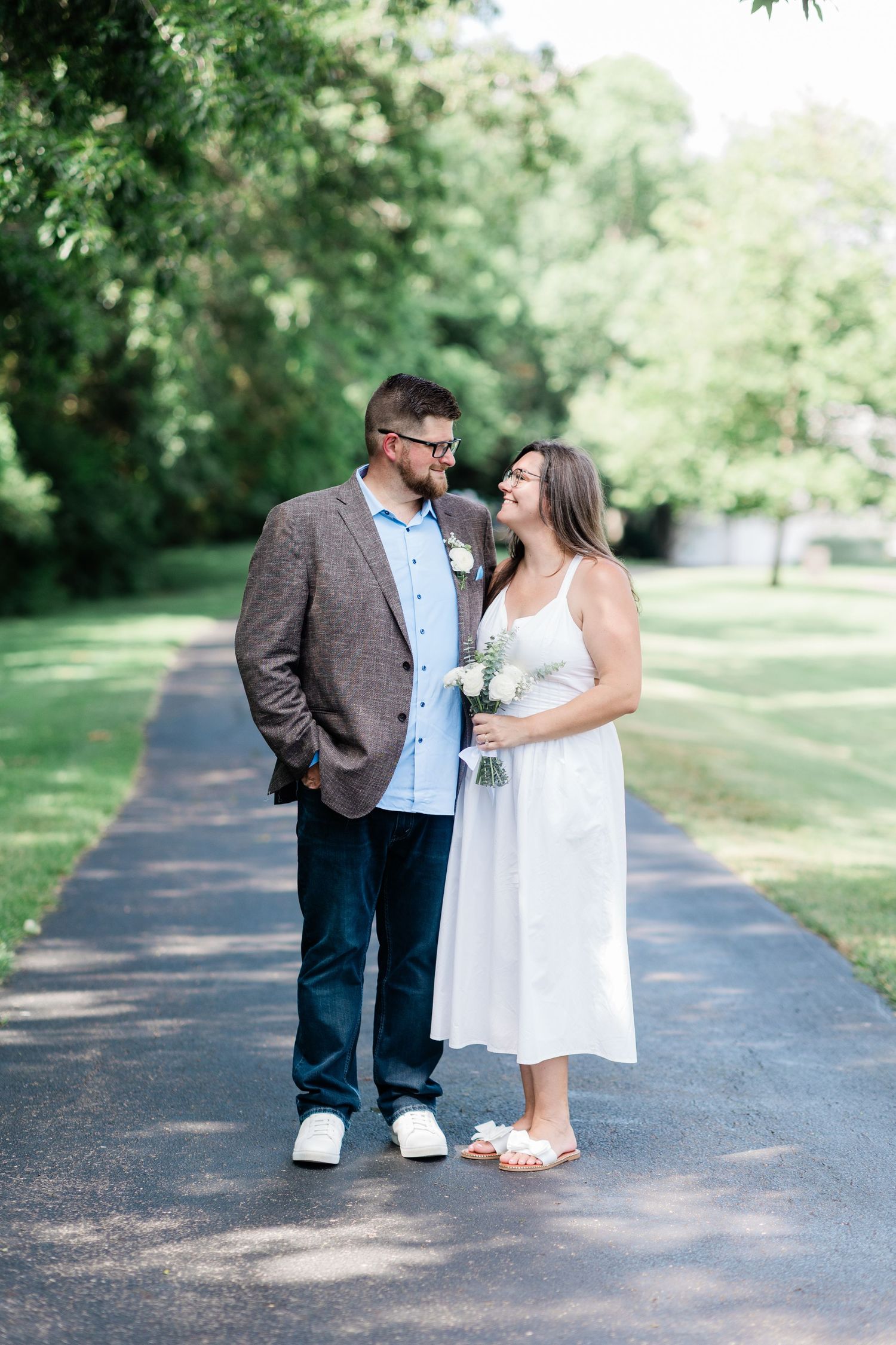 Bride in white dress holding bouquet and groom in brown blazer smiling at each other on a tree-lined pathway.