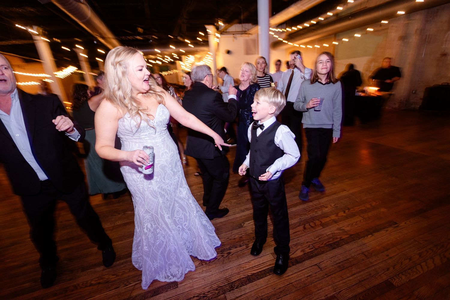 Bride in lavender gown dancing joyfully with a young boy in a vest on a wooden wedding reception dance floor.