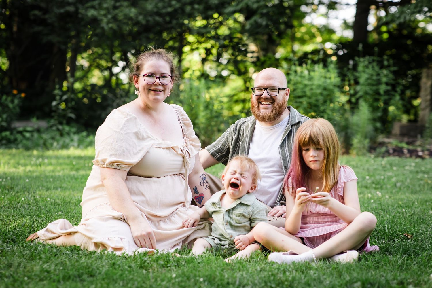 Family of four sitting on grass, mother and father with two young children laughing outdoors.