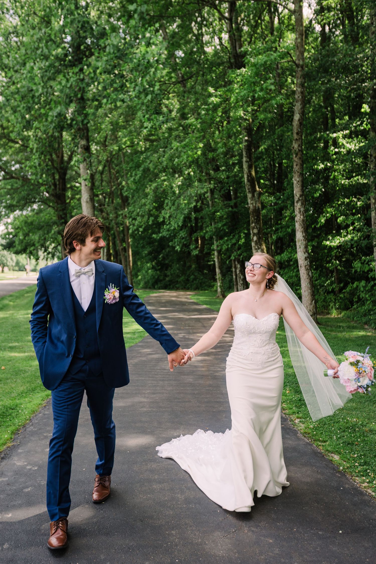 Bride and groom holding hands walking down a scenic tree-lined path on their wedding day.
