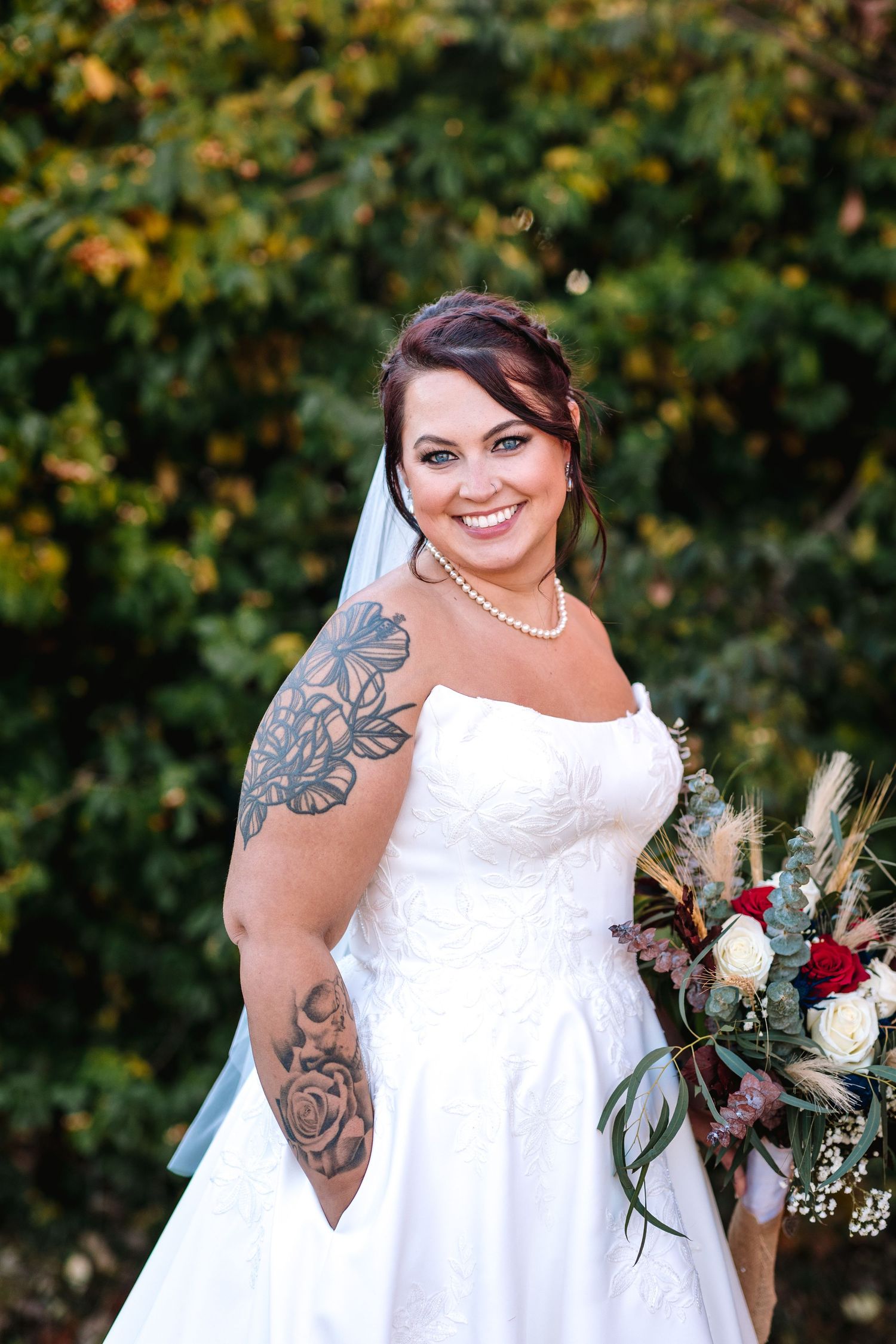 Smiling bride in a strapless white gown with arm tattoos holds a bouquet of red and white flowers.