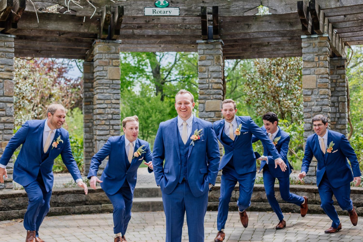 Groom and groomsmen in royal blue suits jumping playfully in a stone garden pavilion.